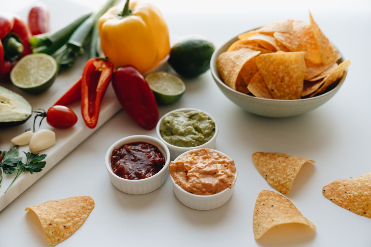 Colorful Mexican cuisine spread with chips, salsa, guacamole, and fresh vegetables on a white background.